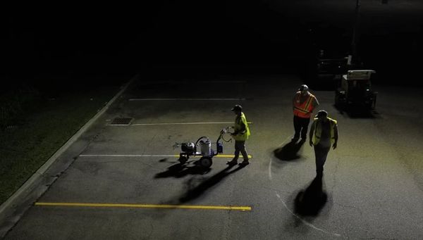 Workers in reflective vests marking parking lot lines at night under bright lights.