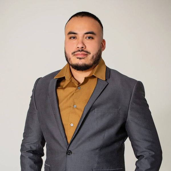 Confident man in a gray blazer and brown shirt posing against a plain background.
