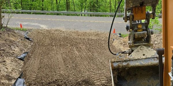 Excavator grading soil near a roadside with trees in the background.