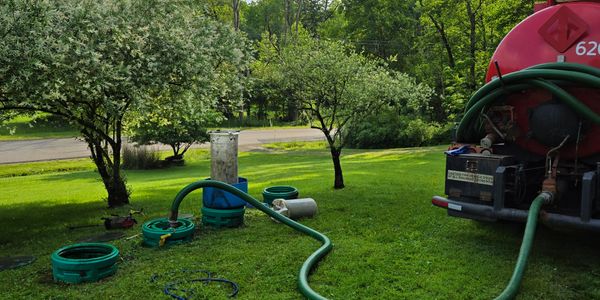 A green hose connected to a red tanker on a grassy lawn with trees in the background.