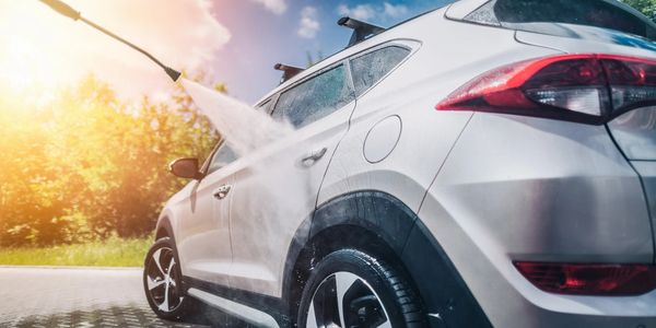 A white SUV being washed with a high-pressure water spray on a sunny day.