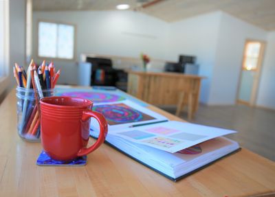 A red mug, colored pencils, and an open book on a wooden table in a bright room.