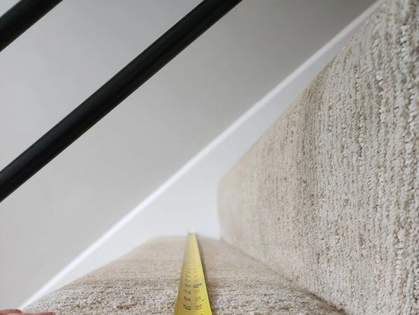Measuring the width of a carpeted stair with a yellow tape measure against a white wall.