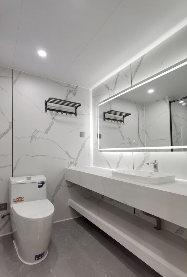Modern bathroom with marble walls, a toilet, and a large lit mirror above a sleek white sink.