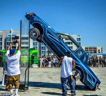 A blue lowrider car performing a hydraulic lift at a car show.