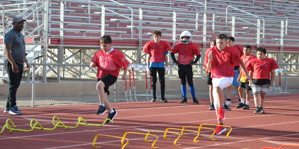 Young boys in red shirts practice agility drills on a track with a coach.