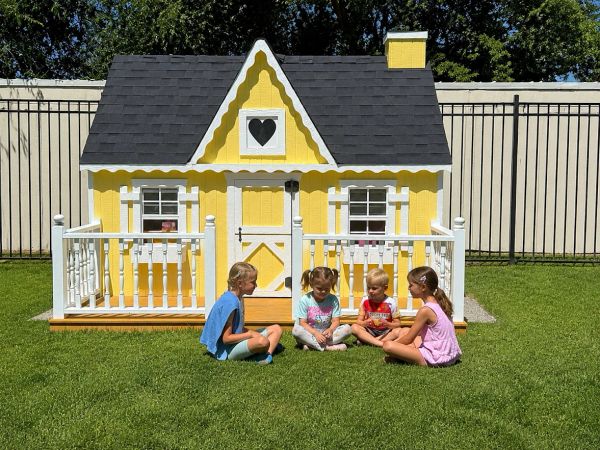 Four children sit in front of a yellow playhouse on green grass.
