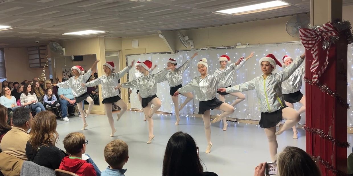 Ballet dancers in silver tops and Santa hats perform for an audience indoors.