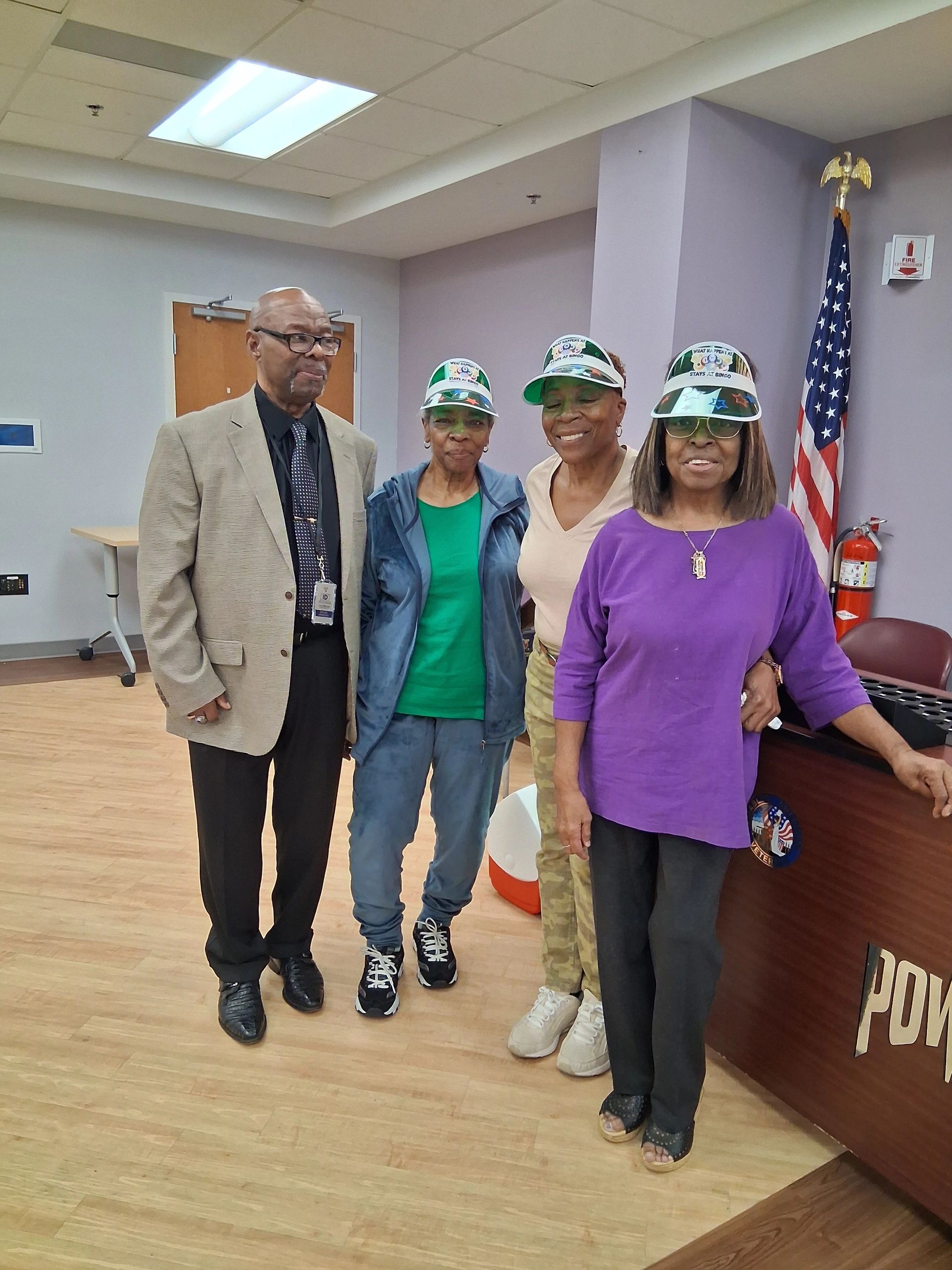 Four seniors posing indoors, three women wearing bingo visors and a man in a suit.