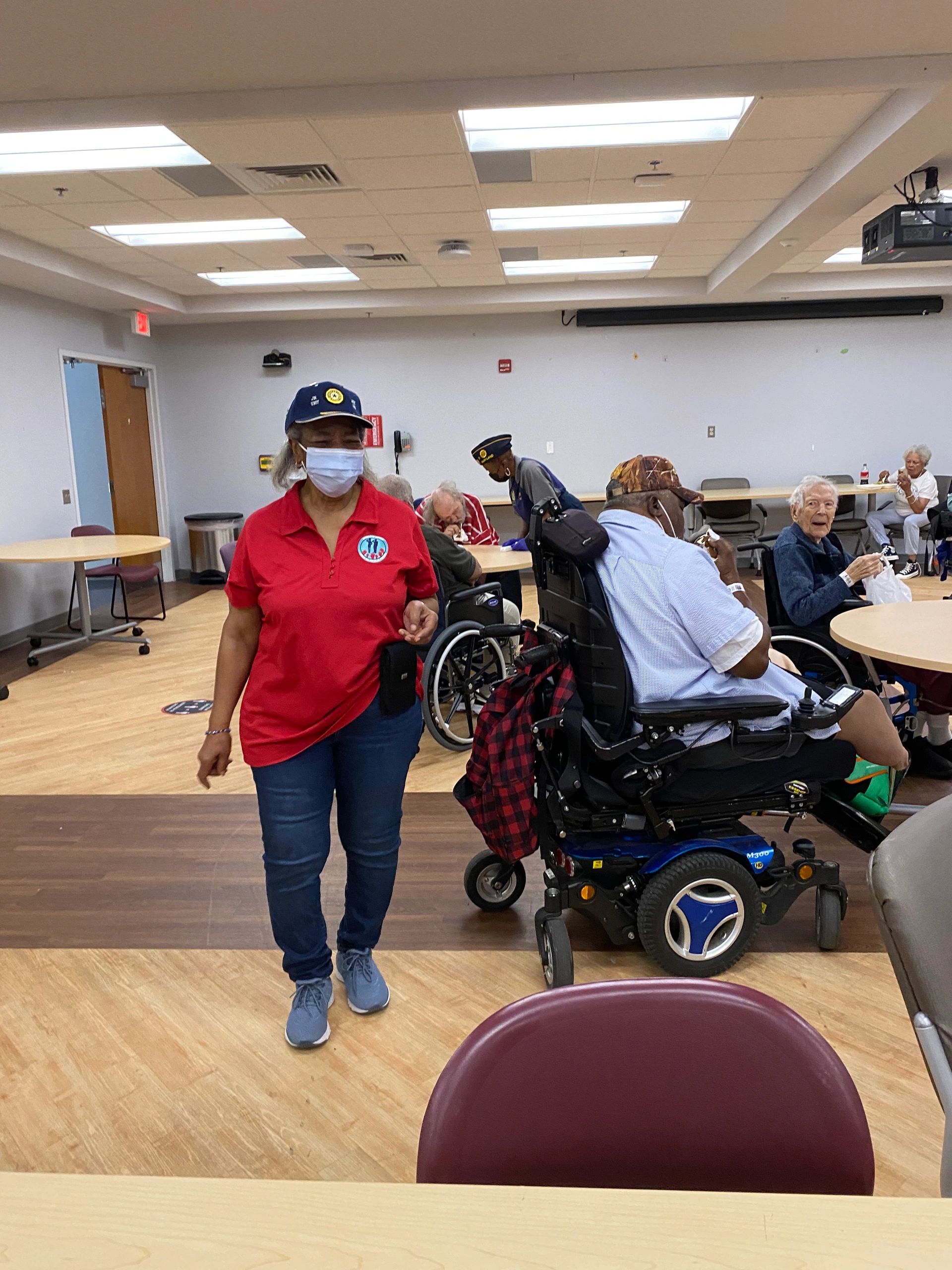 A woman in a mask walks in a room with elderly people, some in wheelchairs.