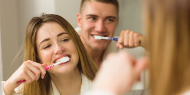 A couple brushing their teeth together in front of a bathroom mirror.