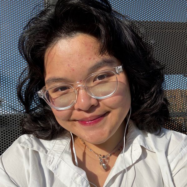 Smiling person with glasses and necklace in sunlight.