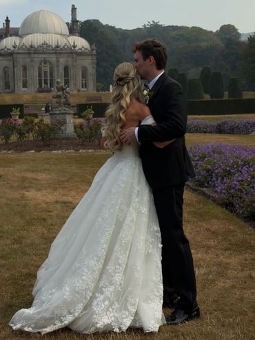 Newlyweds embrace in a garden with historic architecture.
