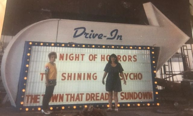 Two people stand in front of a vintage drive-in movie sign listing horror films.