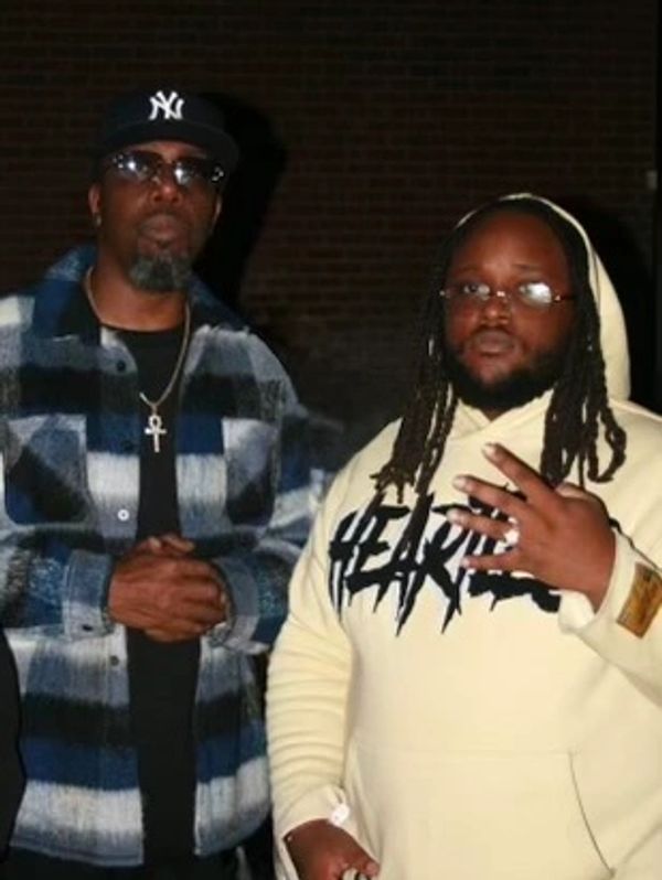 Two men posing together, one in a checkered jacket and the other in a hoodie.