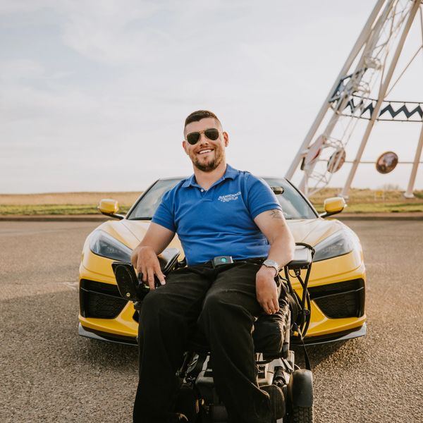 Man in a wheelchair smiling in front of a yellow sports car.