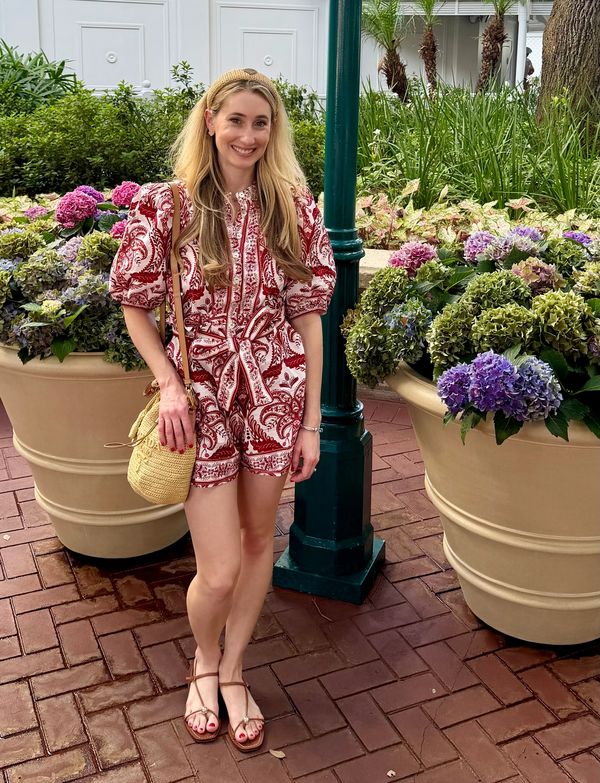 A woman in a red and white romper stands smiling between large flower pots.