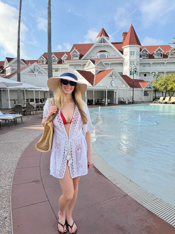 Woman in a white cover-up and sunhat by a resort pool on a sunny day.