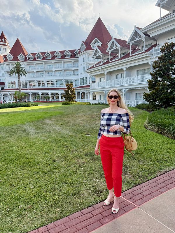 Woman in red pants and checkered top stands on a path near a large white building with red roofs.