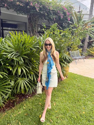 A stylish woman in a white dress and blue scarf walks on grass near lush greenery.
