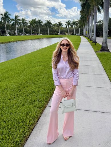 A stylish woman in pastel pink pants and lavender blouse stands on a sidewalk by a pond and palm trees.