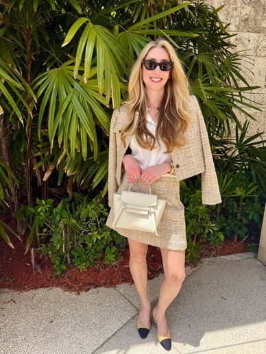 A stylish woman in a beige tweed skirt suit and sunglasses stands smiling outdoors with a white handbag.