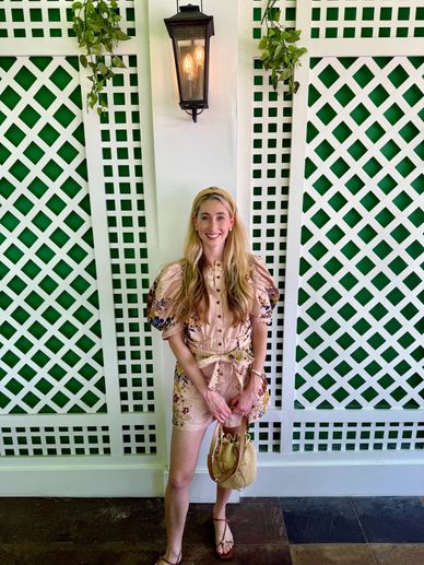 Young woman in floral dress and headband stands smiling against a white lattice wall with hanging plants.