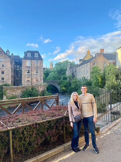 Couple posing by a scenic riverside with historic buildings in the background under a blue sky.