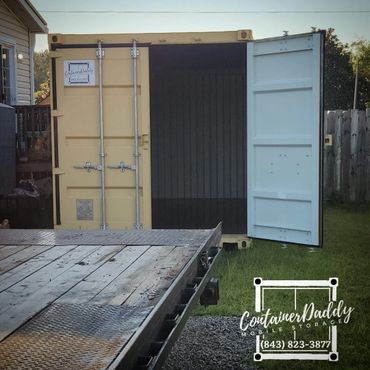 Yellow mobile storage container with open door, parked on grass beside a wooden fence.