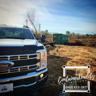 A Ford truck and a green shipping container in a rural field under a clear sky.