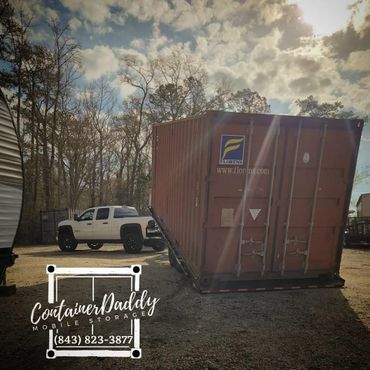 A large shipping container being transported by a white pickup truck on a sunny day.