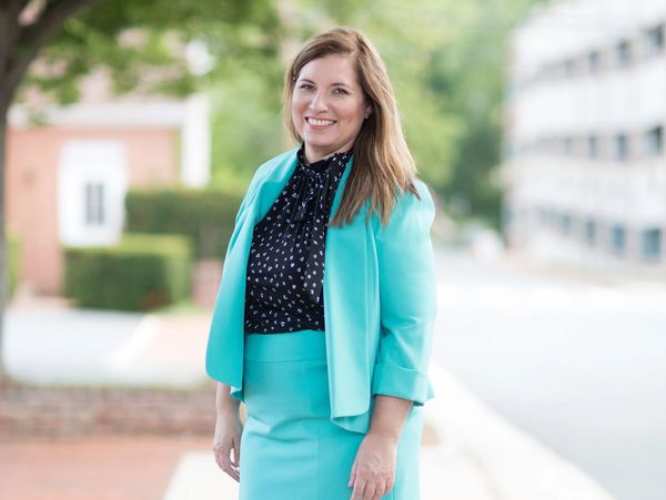 Woman in a turquoise skirt suit smiling outdoors on a brick sidewalk.