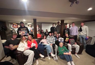 A large group of young adults gathered in a cozy living room, holding gifts and smiling.