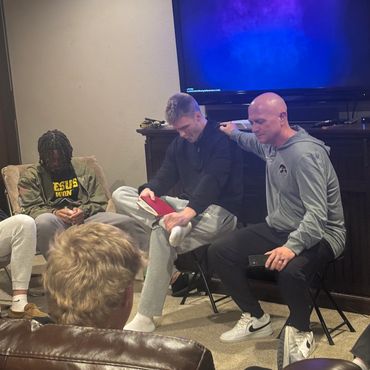 A group of men sitting in a living room, some with heads bowed, one holding a book.