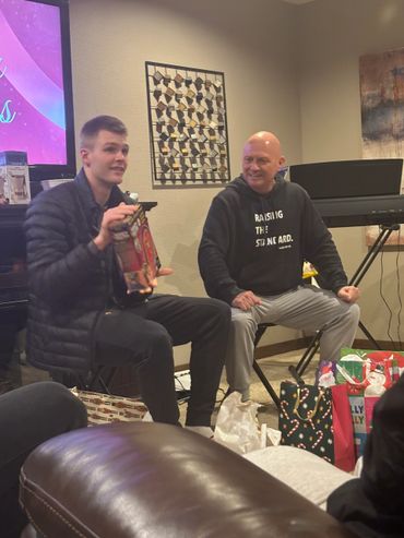 Two men sitting indoors, one holding a boxed gift, surrounded by festive bags.