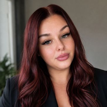 A woman with long, wavy auburn hair and natural makeup poses indoors.