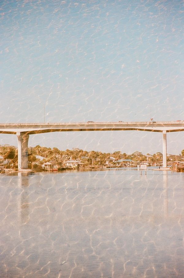 A bridge stretches over calm water with houses and trees in the background.