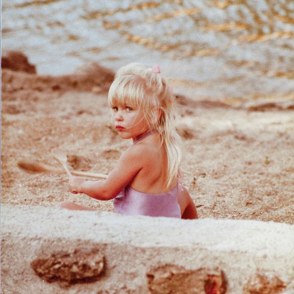 A young girl with blonde hair plays in the sand near water.