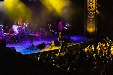 A lively band performs on stage with vibrant yellow lighting and an enthusiastic crowd.