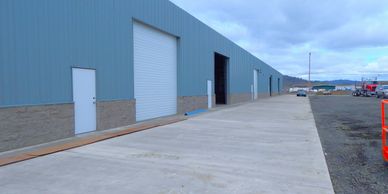 Long exterior view of a blue industrial warehouse with multiple doors and a concrete driveway.