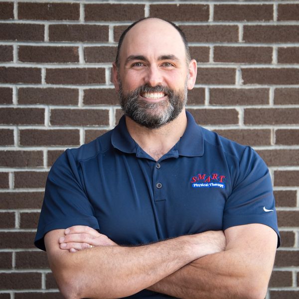 Smiling man with a beard and crossed arms in front of a brick wall.