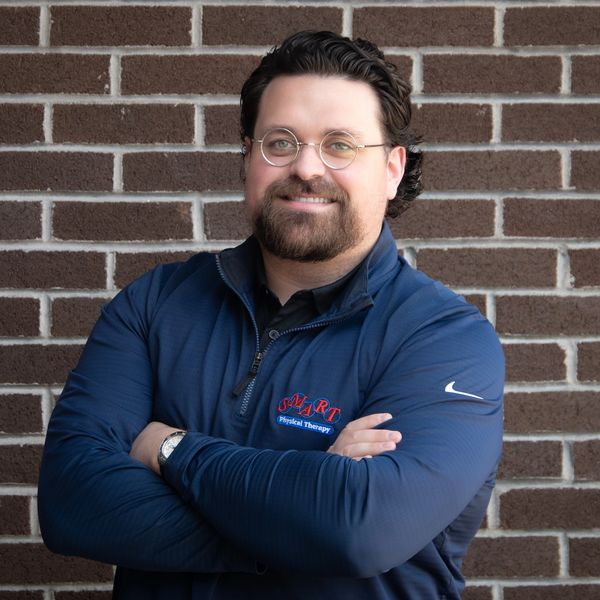 Man in glasses and blue jacket smiling against a brick wall.