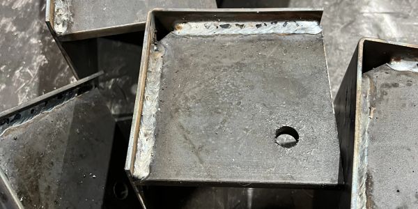 Close-up of welded metal boxes with a drilled hole on one.