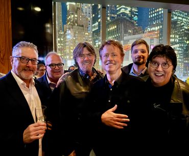 Group of six men posing cheerfully indoors with city lights in the background.