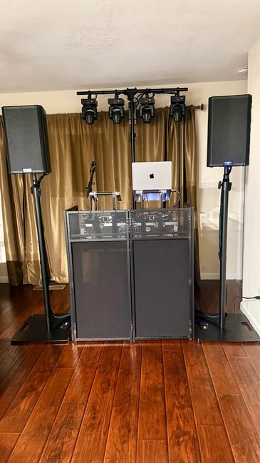 DJ setup with speakers, lighting, and a laptop in a room with wooden floor.