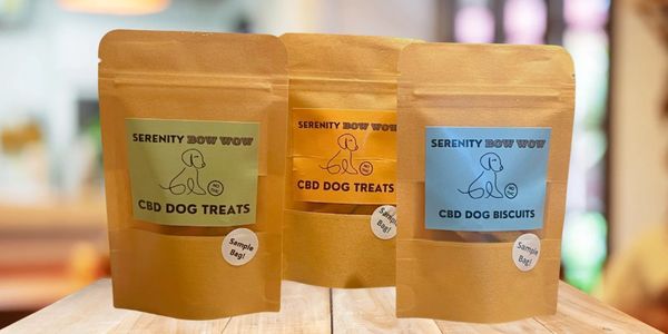 Three sample bags of Serenity Bow Wow CBD dog treats and biscuits on a wooden surface.