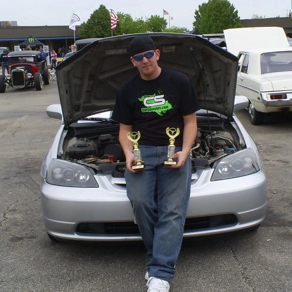 Man leaning on car holding two trophies.