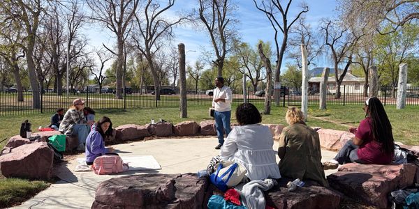 Group of people seated in a circle outdoors on rocks listening to a man standing.