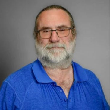 Smiling older man with glasses and a beard in a blue shirt.
