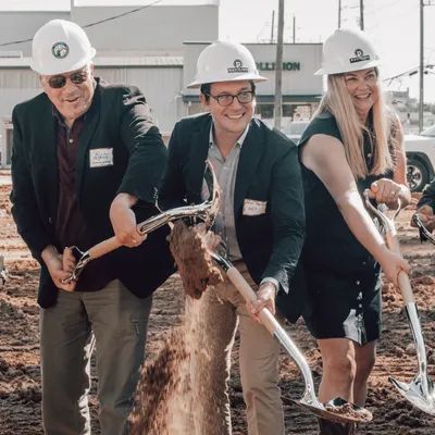Three people in hard hats shovel dirt at a groundbreaking event.